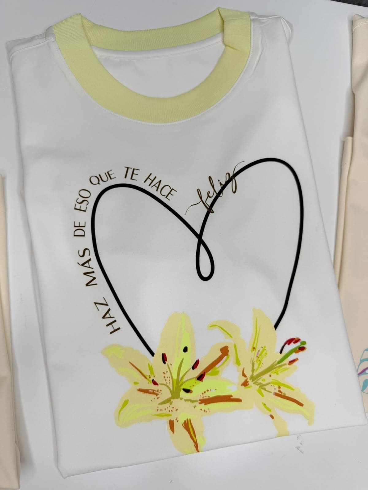 Camiseta blanca con cuello amarillo, corazón, lirios y la frase Haz más de eso que te hace feliz