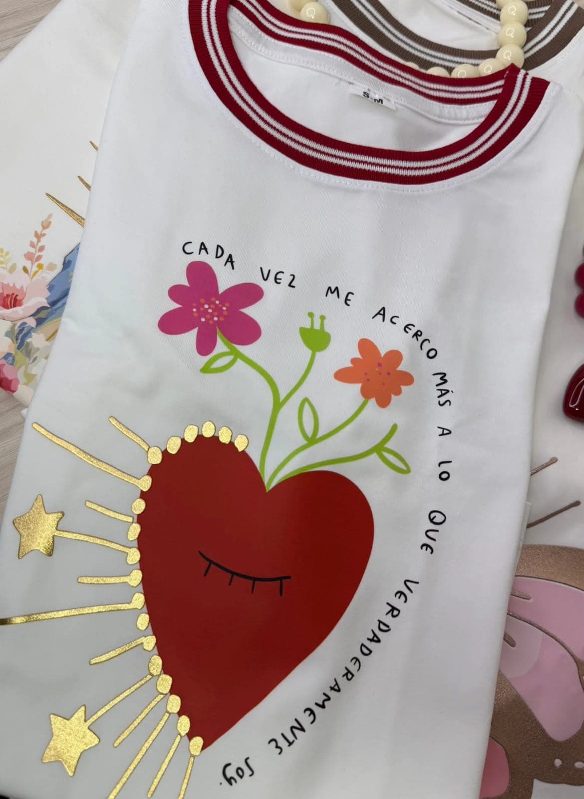 Camiseta blanca con corazón rojo, flores y rayos dorados, cuello tejido a rayas