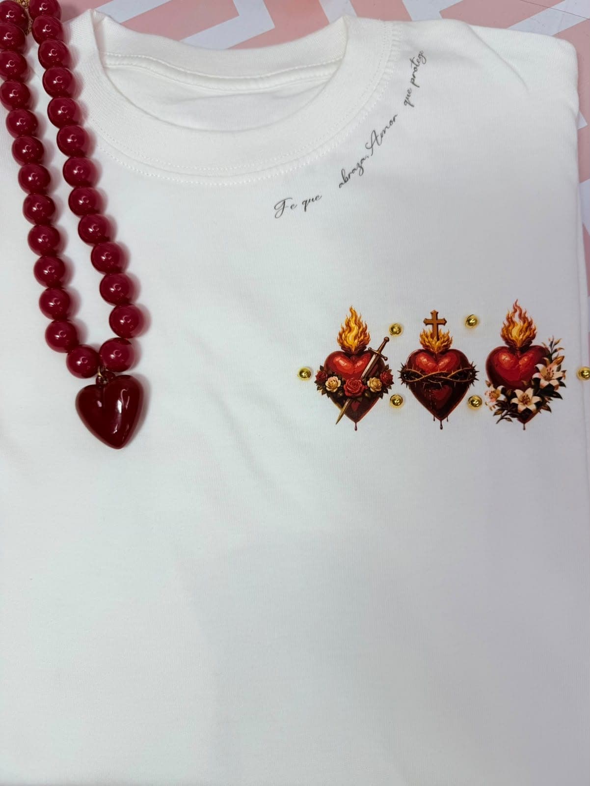 Camiseta crema con tres sagrados corazones y la frase Fe que abraza, amor que protege